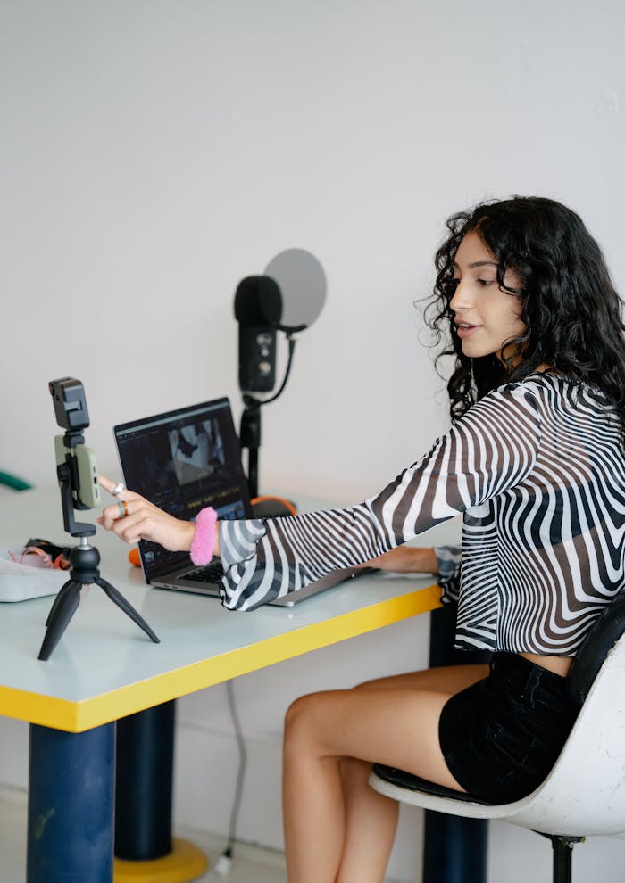 about-002 A woman recording herself with a smartphone and laptop at a desk indoors.