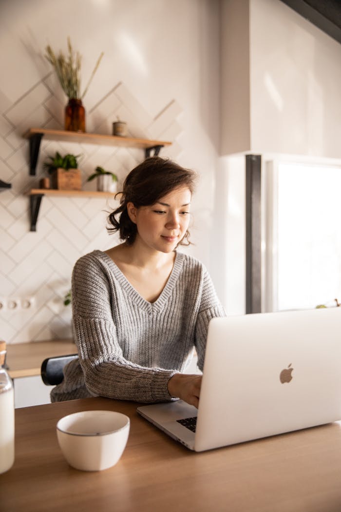 project-01-c Young female in gray sweater sitting at wooden desk with laptop and bottle of milk near white bowl while browsing internet on laptop during free time at home