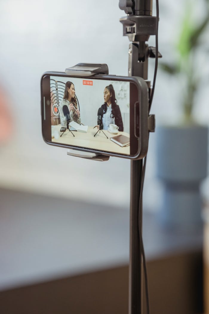project-03-a Young diverse women sitting at table with microphones and gadgets and talking while recording podcast on smartphone placed on tripod in studio